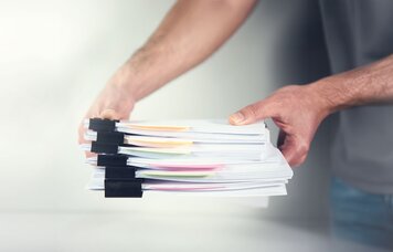 Eine Person legt einen Stapel gebündelter Papiere auf einen Tisch. | © Foto: AdobeStock #414536160 | ARAMYAN 