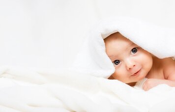 Ein Baby mit blaugrauen Augen liegt in einem weichen Handtuch eingewickelt auf einem Tisch. | © Foto: AdobeStock #171484206 | ballabeyla